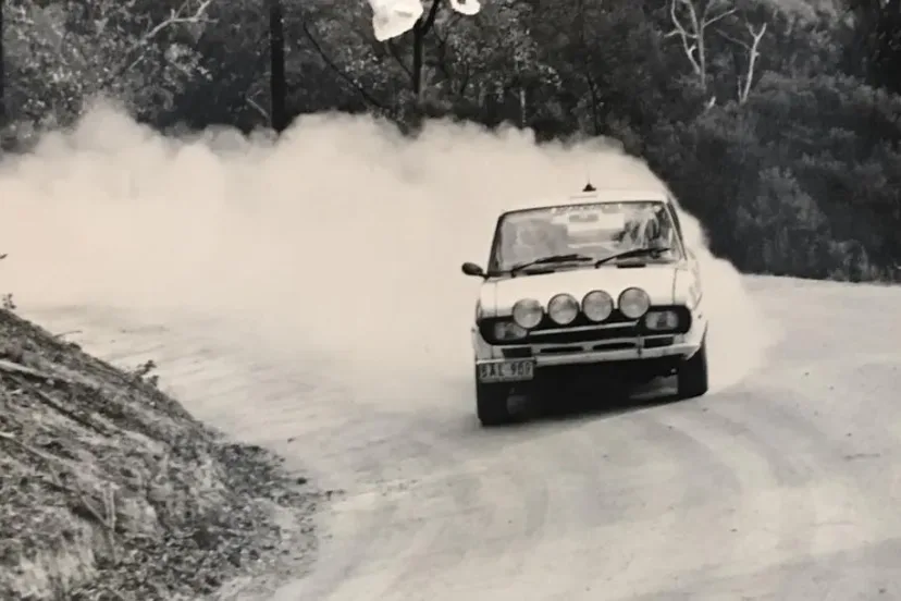 A white Fiat X1/9 rally car trailing a wall of dust on a gravel forest road during the Akademos Rally.