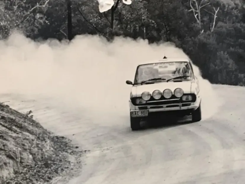 A white Fiat X1/9 rally car trailing a wall of dust on a gravel forest road during the Akademos Rally.