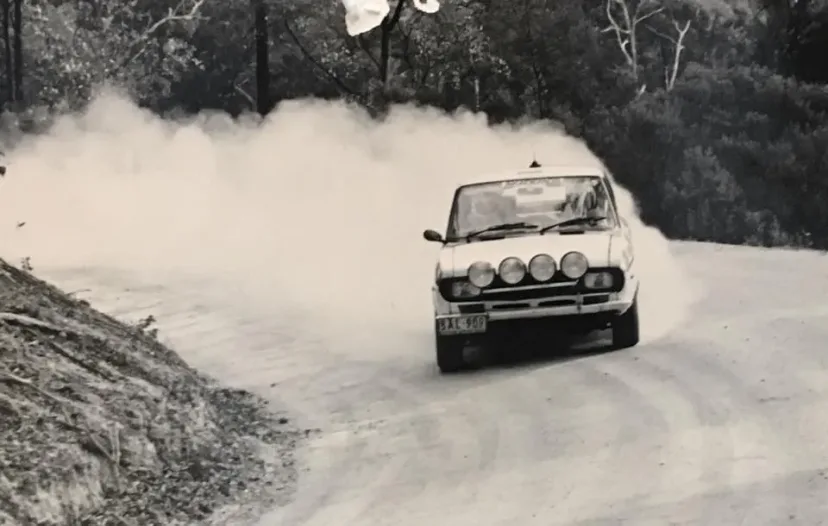 A white Fiat X1/9 rally car trailing a wall of dust on a gravel forest road during the Akademos Rally.