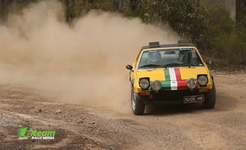 A yellow Fiat X1/9 rally car emerging from its own dust cloud on a gravel stage.