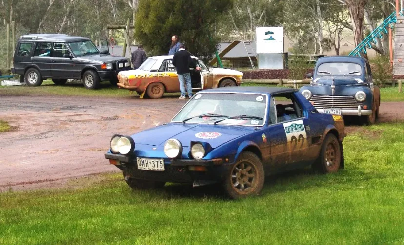 A blue Fiat X1/9 on grass at the Ye Olde BP Rally, with a Land Rover and vintage cars behind.